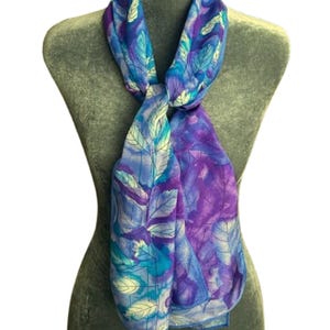 May include: A lightweight scarf featuring a vibrant watercolor-inspired pattern of purple and blue hues. The design is adorned with delicate, outlined leaf motifs in a soft cream color, creating a layered botanical effect across the fabric.