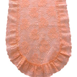 May include: A peach-colored lace table runner featuring a repeating floral pattern throughout the center panel. The runner is finished with a gathered, ruffled lace border that follows the rounded edge of the fabric, creating a textured look.