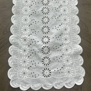May include: A white rectangular table runner featuring intricate eyelet embroidery with a repeating floral pattern. The edges are finished with a scalloped border, and the fabric displays a series of cut-out daisy-like flowers and vine motifs.