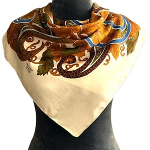 May include: A square scarf featuring a cream-colored base with a vibrant border of autumn-toned leaves in shades of orange, brown, and green, accented by blue ribbons and dark brown paisley patterns. The text "Made in England Liberty of London" is visible.