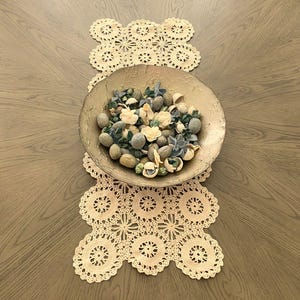 May include: A decorative centerpiece featuring a gold-toned bowl filled with assorted potpourri, resting on a cream-colored, crocheted table runner. The runner has a floral design. The table is made of wood.