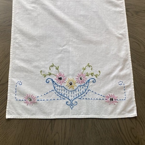 May include: White embroidered table runner with a floral design. The embroidery features pink, yellow, and black flowers, green leaves, and blue accents. The runner is made of a textured fabric.