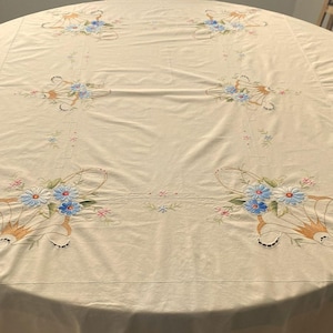 May include: A white tablecloth with embroidered floral designs in shades of blue, white, and pink. The tablecloth features a repeating pattern of stylized floral bouquets with delicate leaves and stems.