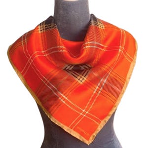 May include: A vibrant orange square scarf with a classic plaid pattern in white, black, and brown. The scarf has a gold border and the word "GODELLO" is printed on the corner. The scarf is displayed on a mannequin.