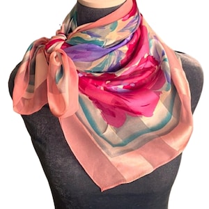 May include: A square silk scarf with a floral design. The scarf features a vibrant color palette of pink, purple, and teal, with a peach border. The scarf is knotted at the neck, showcasing the delicate, sheer fabric.