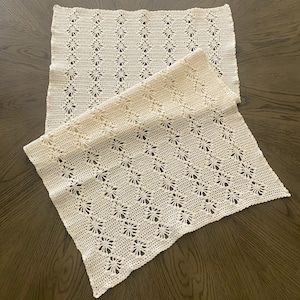 May include: Two off-white, crocheted placemats with a floral pattern. The placemats are rectangular and have a delicate, openwork design. The crochet work is detailed, with each flower carefully crafted. The placemats are placed on a dark wooden surface.