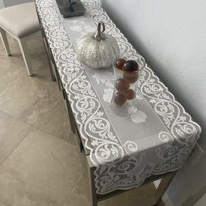 May include: A gray table runner with a white floral lace design and a decorative silver pumpkin. A clear glass vase holds several brown decorative spheres. The runner is on a gray cabinet.