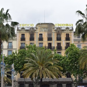 May include: A yellow building with the words "Edificio Condeminas" on the front. The building is surrounded by palm trees and other greenery.