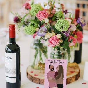 May include: A pink table number card with the text "Table Two" and an illustration of a couple embracing. The card is on a wooden stand and surrounded by pink and white flowers in a glass vase.