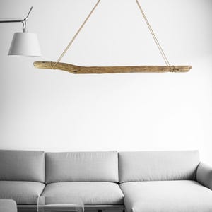 Peut inclure: Un luminaire suspendu en bois flotté, suspendu par deux cordes. Le bois est de couleur brun clair et a une texture rugueuse. Les cordes sont de couleur beige clair. La lumière est au-dessus d'un canapé gris.