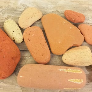 May include: A selection of orange and tan stones of different sizes and shapes. One stone has a jute string attached. The stones are displayed on a light-coloured, textured surface. The stones vary in shade.