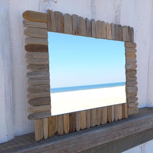 May include: Rectangular mirror with a frame made of weathered driftwood pieces. The mirror reflects a beach scene with a blue sky and ocean. The driftwood frame has a natural, rustic appearance, adding a coastal aesthetic.