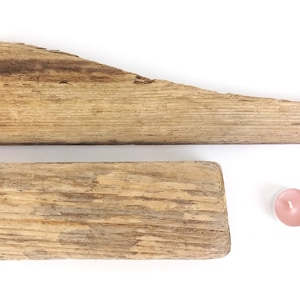 Peut inclure: Deux morceaux de bois flotté vieilli, l'un long et étroit, l'autre plus court et plus large, disposés sur un fond blanc. Une petite bougie rose se trouve à droite du bois.