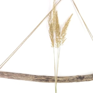 May include: A rustic wall hanging featuring a curved piece of driftwood suspended by twine. Dried wheat stalks are positioned in the center. The natural wood has a weathered appearance, and the twine is a light beige color.