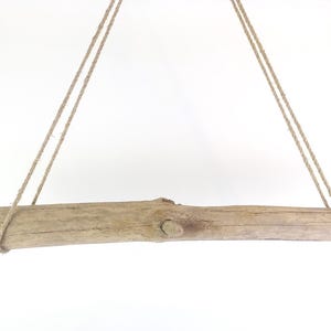 Op de afbeelding: Een natuurlijke houten tak, ongeveer 50 cm lang, hangt aan twee touwtjes aan het plafond. De tak is lichtbruin van kleur en heeft een ruwe, natuurlijke textuur.