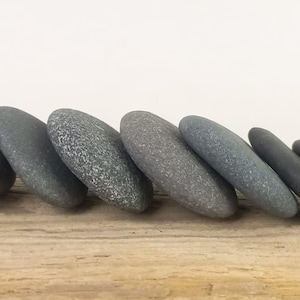 May include: A horizontal row of twelve smooth, dark gray river stones arranged in a line. Each stone has a rounded, flattened shape with a matte texture, varying slightly in size and shade, positioned side by side in a sequential formation.