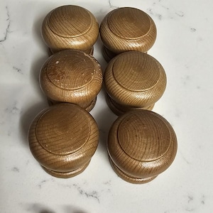 May include: Six wooden knobs with a light brown finish. The knobs have a rounded top and a slightly raised edge. The knobs are arranged in two rows of three. There are also six silver screws included.