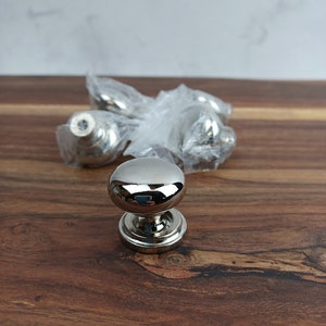 May include: A close-up of a silver cabinet knob with a round, smooth surface. The knob is sitting on a wooden surface.