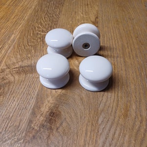 May include: Five white ceramic cabinet knobs with a smooth, glossy finish. The knobs are round and have a slight indentation in the centre.