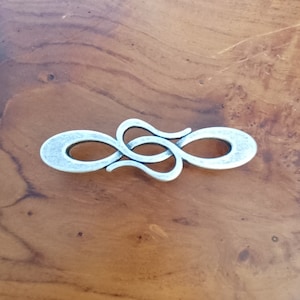 May include: A silver-toned metal clasp featuring an elongated, symmetrical infinity-like design with a textured, matte finish. The hardware consists of two interlocking, curved S-shaped components that form a continuous, horizontal decorative piece.