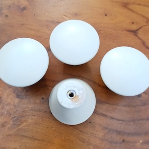 May include: Four white ceramic knobs with a smooth, rounded surface. Each knob has a small hole in the centre for attaching to a drawer or cabinet.