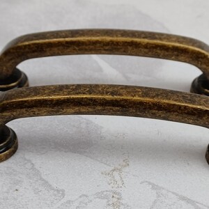 May include: Two antique gold-toned cabinet pulls with a rounded rectangular shape. The pulls have a slightly distressed finish.