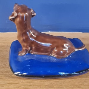 Puede incluir: Una figurita de dachshund de cer&aacute;mica marr&oacute;n sentada sobre una almohada de cer&aacute;mica azul. El dachshund est&aacute; mirando hacia arriba y tiene la cola enroscada alrededor de su cuerpo.