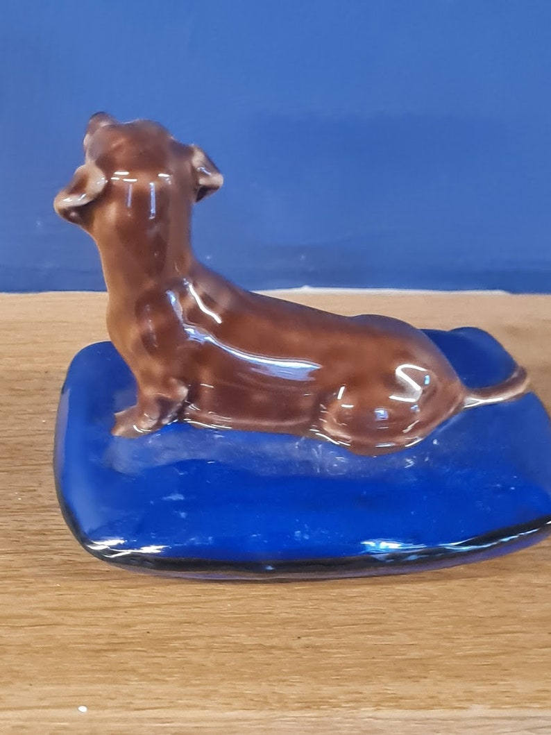 Puede incluir: Una figurita de dachshund de cer&aacute;mica marr&oacute;n sentada sobre una almohada de cer&aacute;mica azul. El dachshund est&aacute; mirando hacia arriba y tiene la cola enroscada alrededor de su cuerpo.