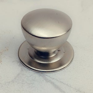 May include: A silver knob with a round top and a flat base. The knob is made of metal and has a brushed finish.
