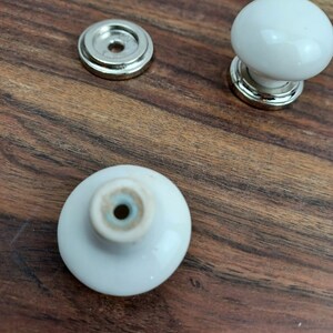 May include: Three white ceramic cabinet knobs with silver metal bases. The knobs are round and have a smooth finish. The bases have a decorative edge.