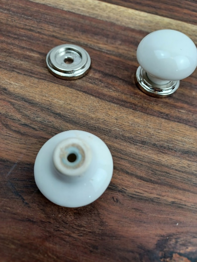 May include: Three white ceramic cabinet knobs with silver metal bases. The knobs are round and have a smooth finish. The bases have a decorative edge.