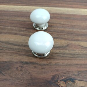 May include: Two white ceramic cabinet knobs with silver metal bases. The knobs are round and have a smooth, glossy finish.