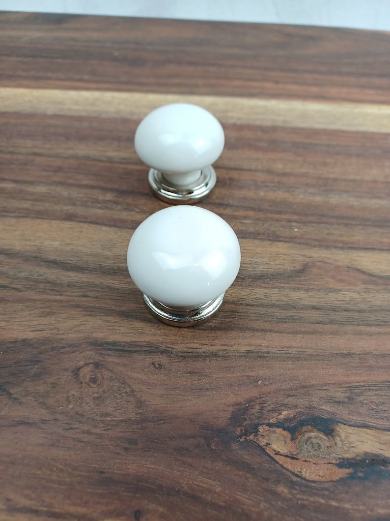 May include: Two white ceramic cabinet knobs with silver metal bases. The knobs are round and have a smooth, glossy finish.