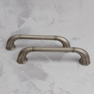 May include: Two antique-style cabinet pulls with a weathered, silver finish. Each pull features a curved handle with decorative detailing and a central cylindrical accent. The pulls are designed for easy installation and add a vintage touch to furniture.
