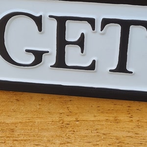 May include: A white sign with black lettering that reads "GET" with a black border. The sign is mounted on a wooden surface.