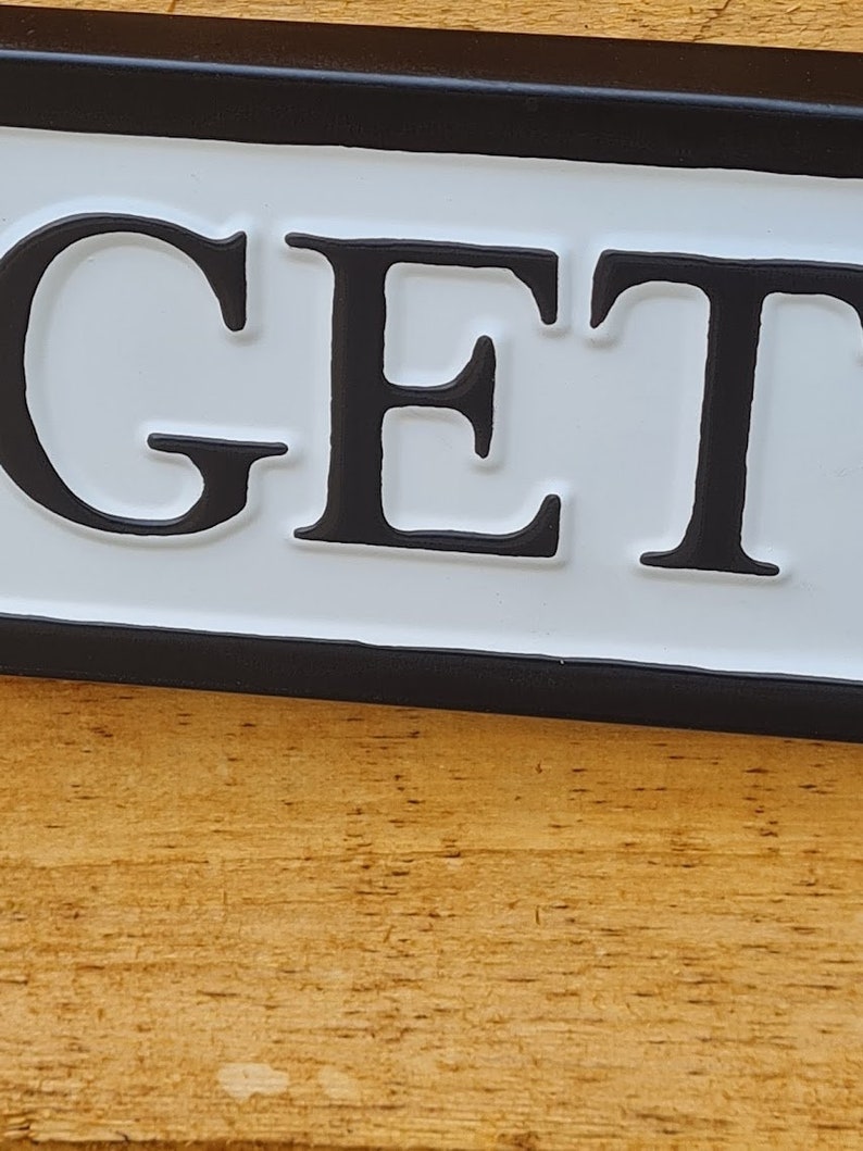 May include: A white sign with black lettering that reads "GET" with a black border. The sign is mounted on a wooden surface.