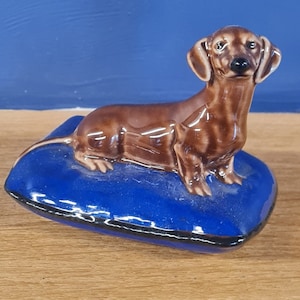 Puede incluir: Una figurita de dachshund de cer&aacute;mica marr&oacute;n sentada en una almohada de cer&aacute;mica azul. El dachshund tiene la nariz marr&oacute;n y negra y los ojos negros.