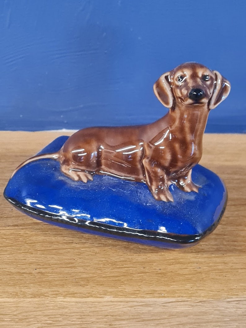 Puede incluir: Una figurita de dachshund de cer&aacute;mica marr&oacute;n sentada en una almohada de cer&aacute;mica azul. El dachshund tiene la nariz marr&oacute;n y negra y los ojos negros.