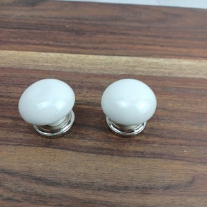 May include: Two white ceramic cabinet knobs with a silver base. The knobs are round and have a smooth finish.