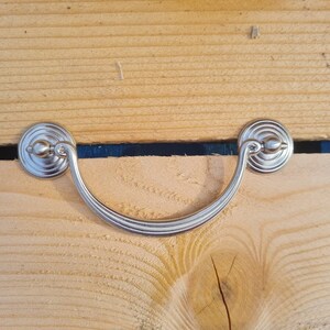 May include: A silver drawer pull with a curved handle and circular backplates. The hardware is mounted on a light-coloured wooden surface with visible grain and a dark blue accent line.