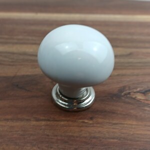 May include: White ceramic cabinet knob with a silver base. The knob is round and has a smooth finish.