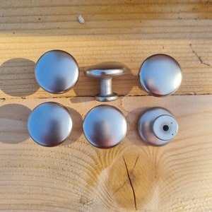 May include: Six silver cabinet knobs, five are round and one is a round knob with a post. The knobs are on a wooden surface.
