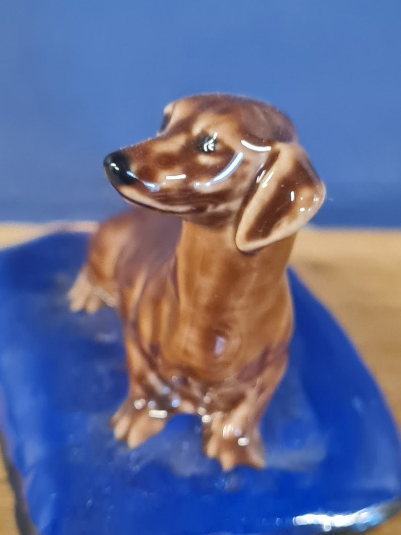 Puede incluir: Una figurita de dachshund marr&oacute;n y blanco sentada sobre una base azul. La figurita est&aacute; hecha de cer&aacute;mica y tiene un acabado brillante.