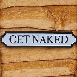 May include: A white sign with black trim and the text "GET NAKED" on a wooden background.