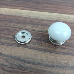 May include: A white ceramic knob with a silver base and a matching silver base plate. The knob is round and has a smooth finish.
