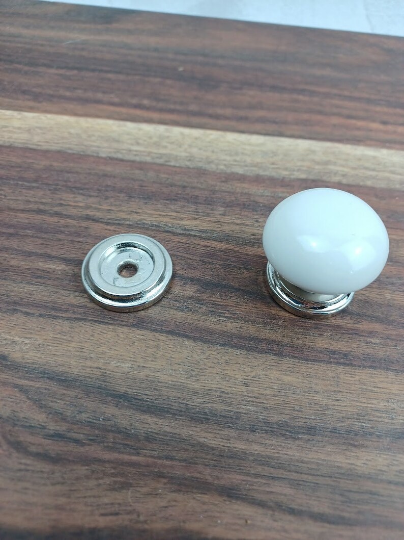 May include: A white ceramic knob with a silver base and a matching silver base plate. The knob is round and has a smooth finish.