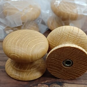 May include: Close-up of four wooden knobs with a natural finish. The knobs have a round top and a cylindrical base. The knobs are packaged in clear plastic bags.