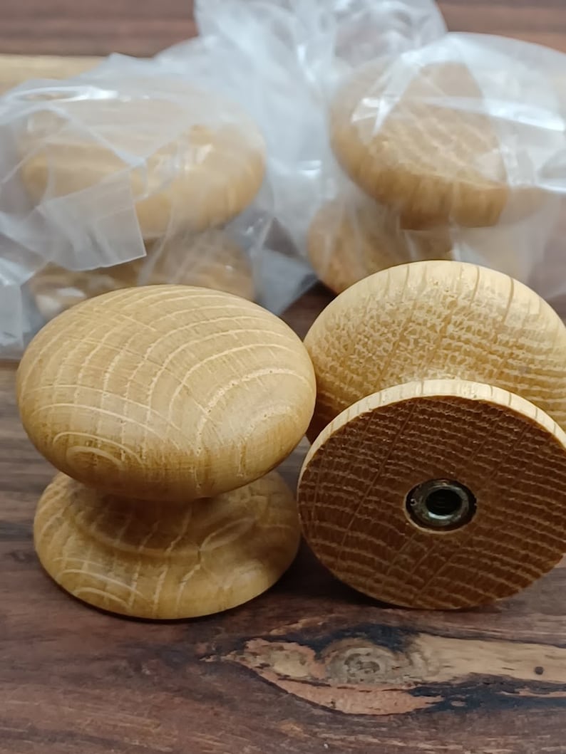 May include: Close-up of four wooden knobs with a natural finish. The knobs have a round top and a cylindrical base. The knobs are packaged in clear plastic bags.
