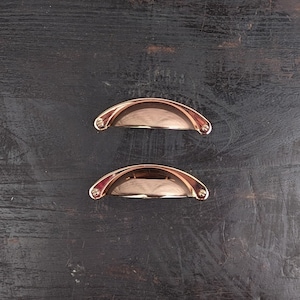 May include: Two small, polished, copper-coloured drawer pulls with a rounded, arched shape. The pulls are set against a dark wood background.