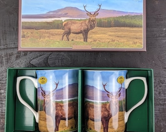 May include: A set of two white ceramic mugs featuring a stag design. The mugs depict a red stag with antlers against a backdrop of a mountain and lake. The mugs are presented in a brown box with a stag image and the text "Stag - Set of Two Mugs".